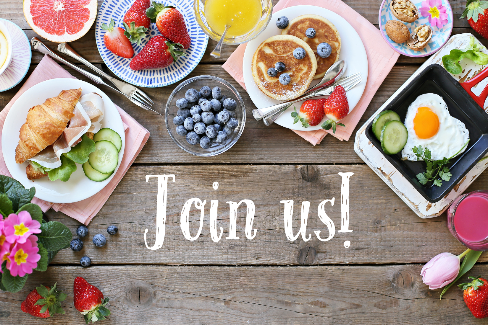 A variety of breakfast foods on wooden table with 'Join Us' in white writing