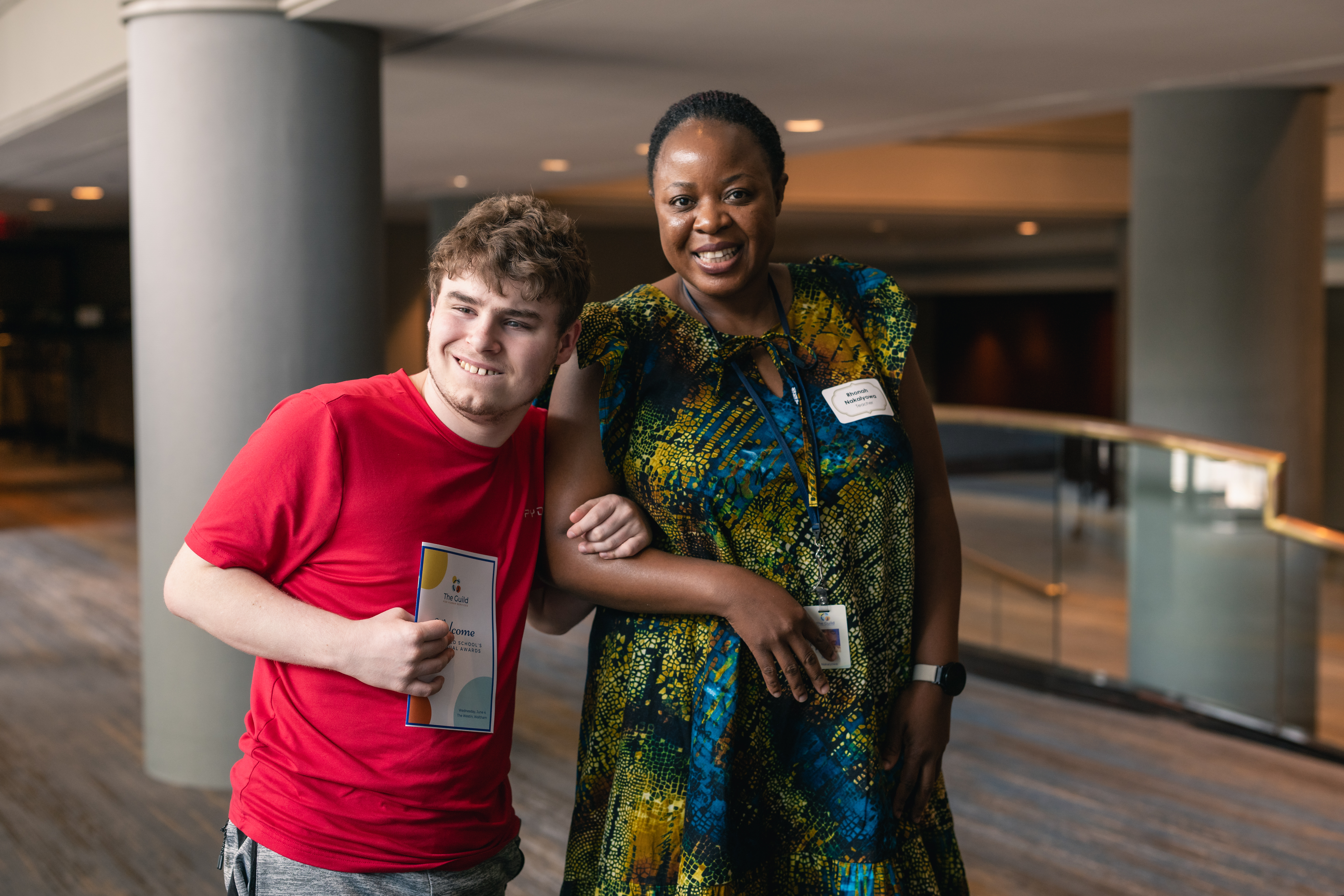 A student stands arm-in-arm with his teacher smiling at the camera.