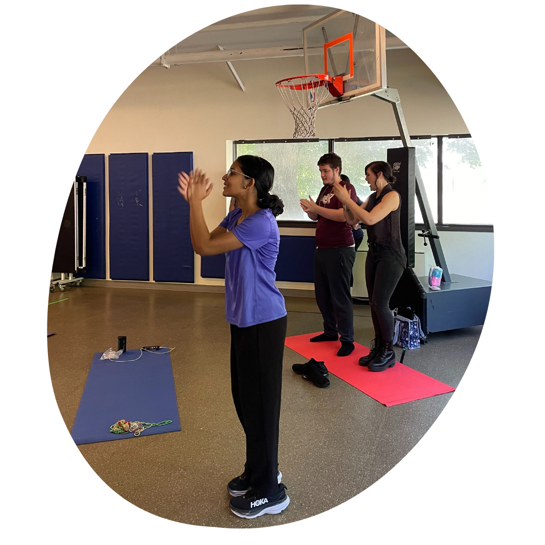Akila Muthukumar stands to the side of a yoga mat and crosses one arm over the other. A student and teacher can be seen following her movements in the background.