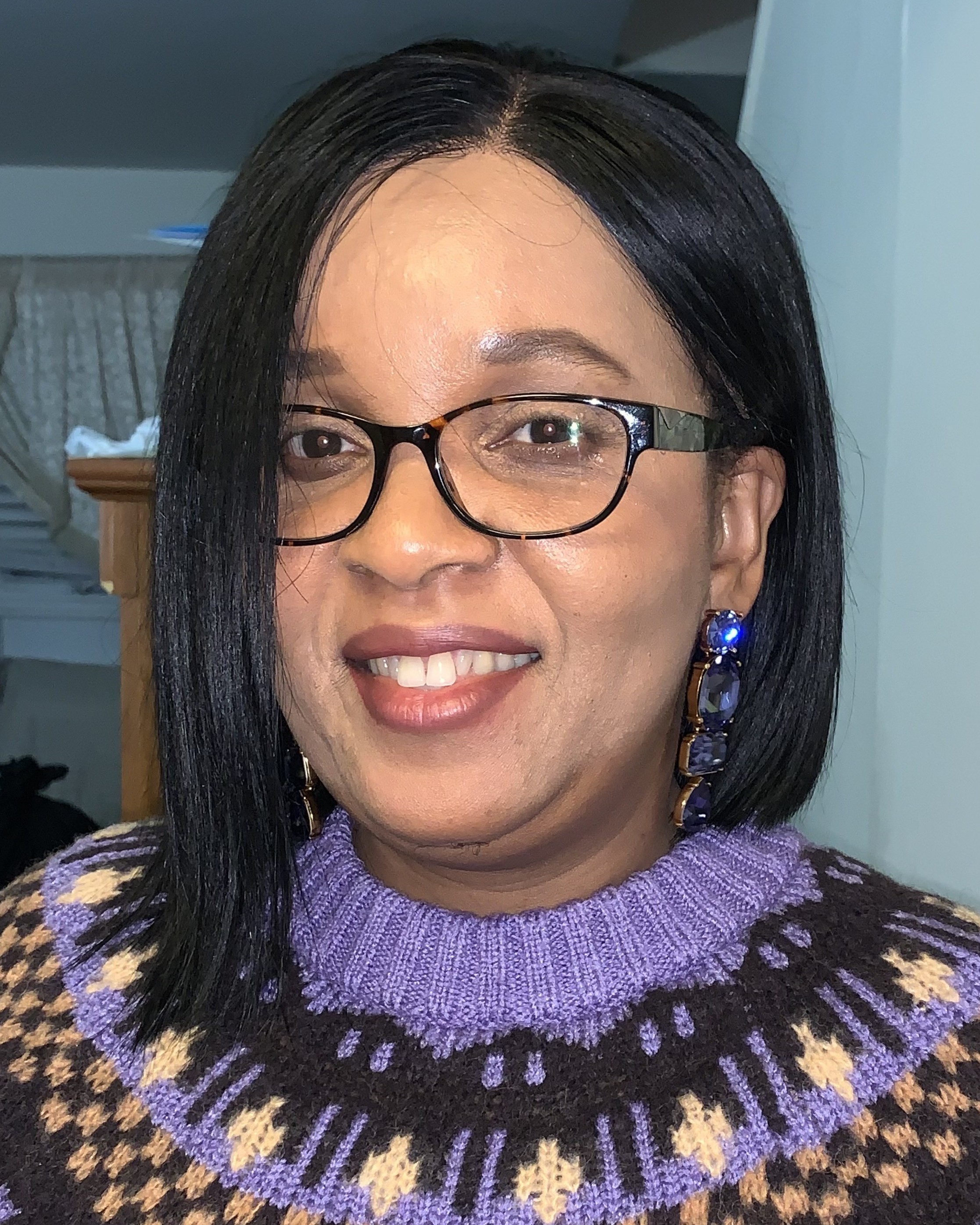 Nerline wears a purple and black woven sweater and glasses and smiles into the camera.