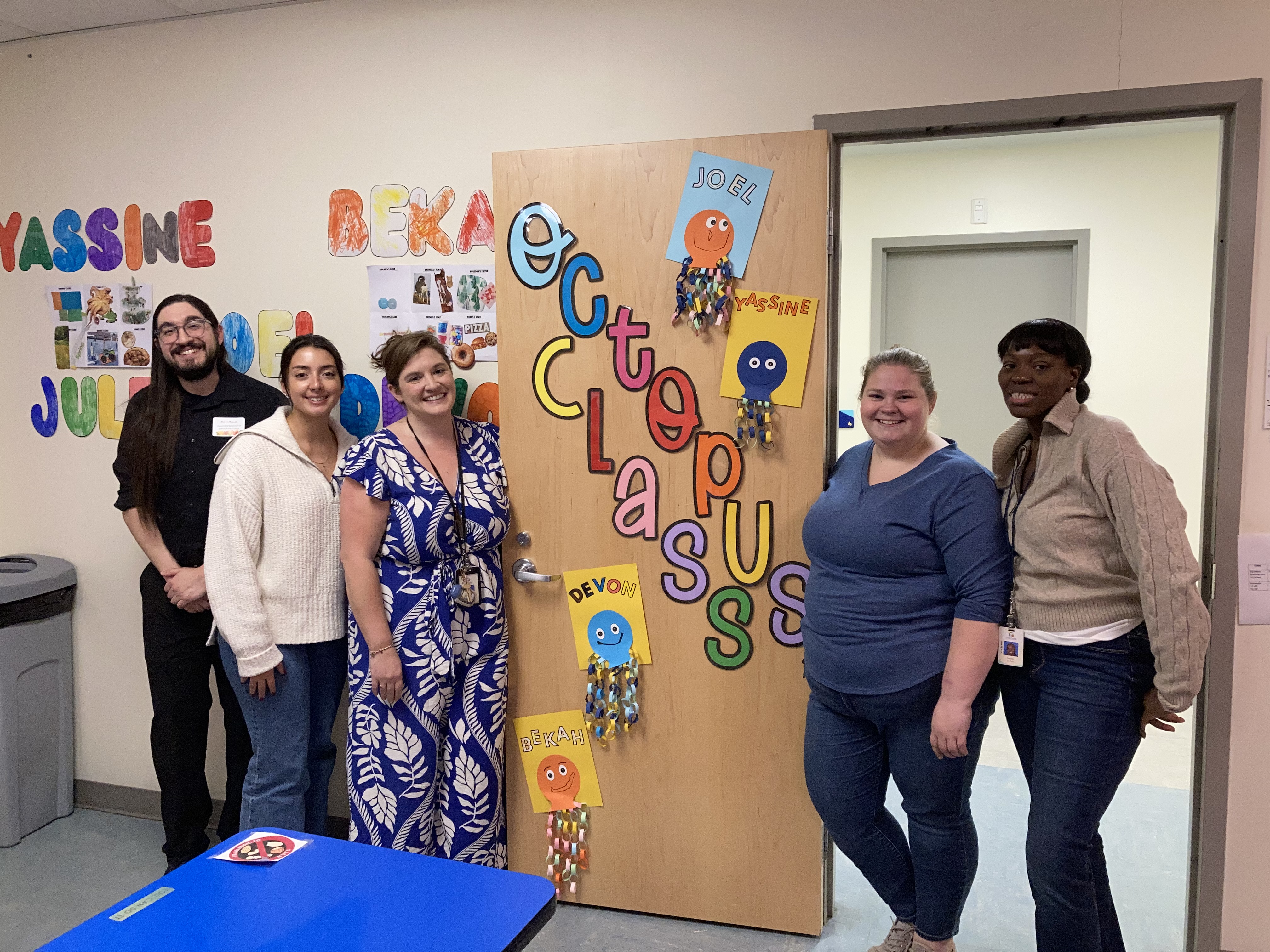 A group of 6 school staff stand in a doorway, which is decorated with colorful letters spelling "Octopus Class".