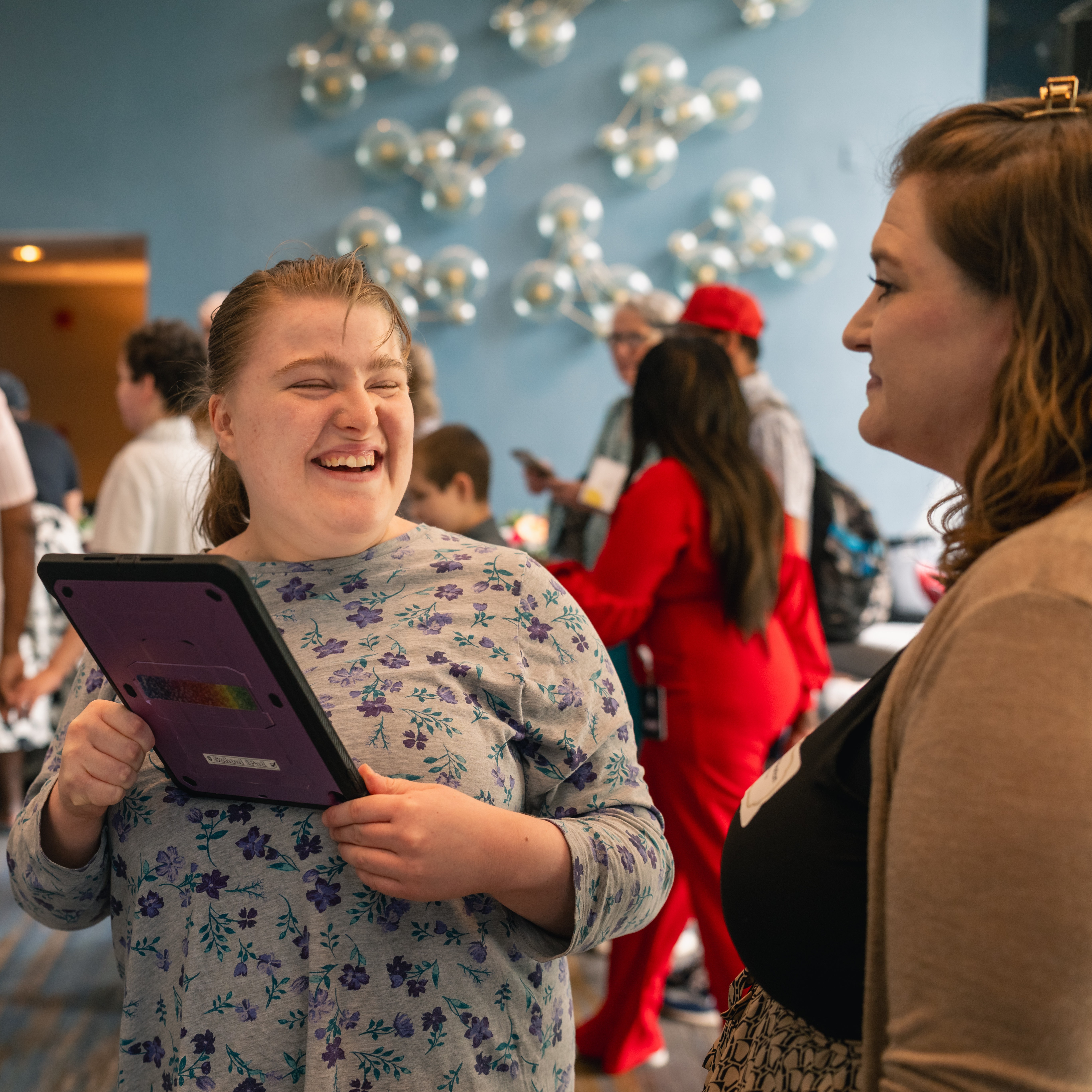 A student smiles at a staff member and shows her something on her communication device.