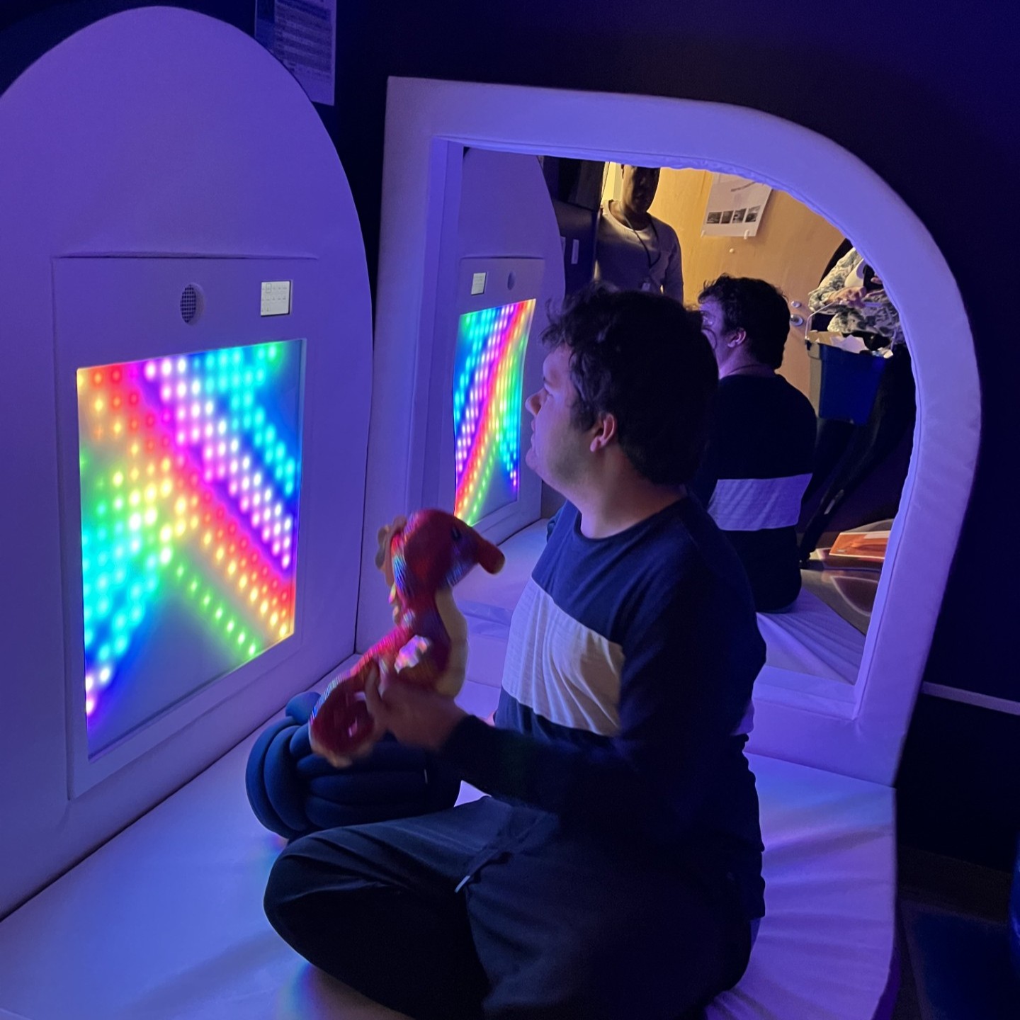 A male student sits on a padded mat in a darkened room and looks at a panel on the wall with multicolored lights.
