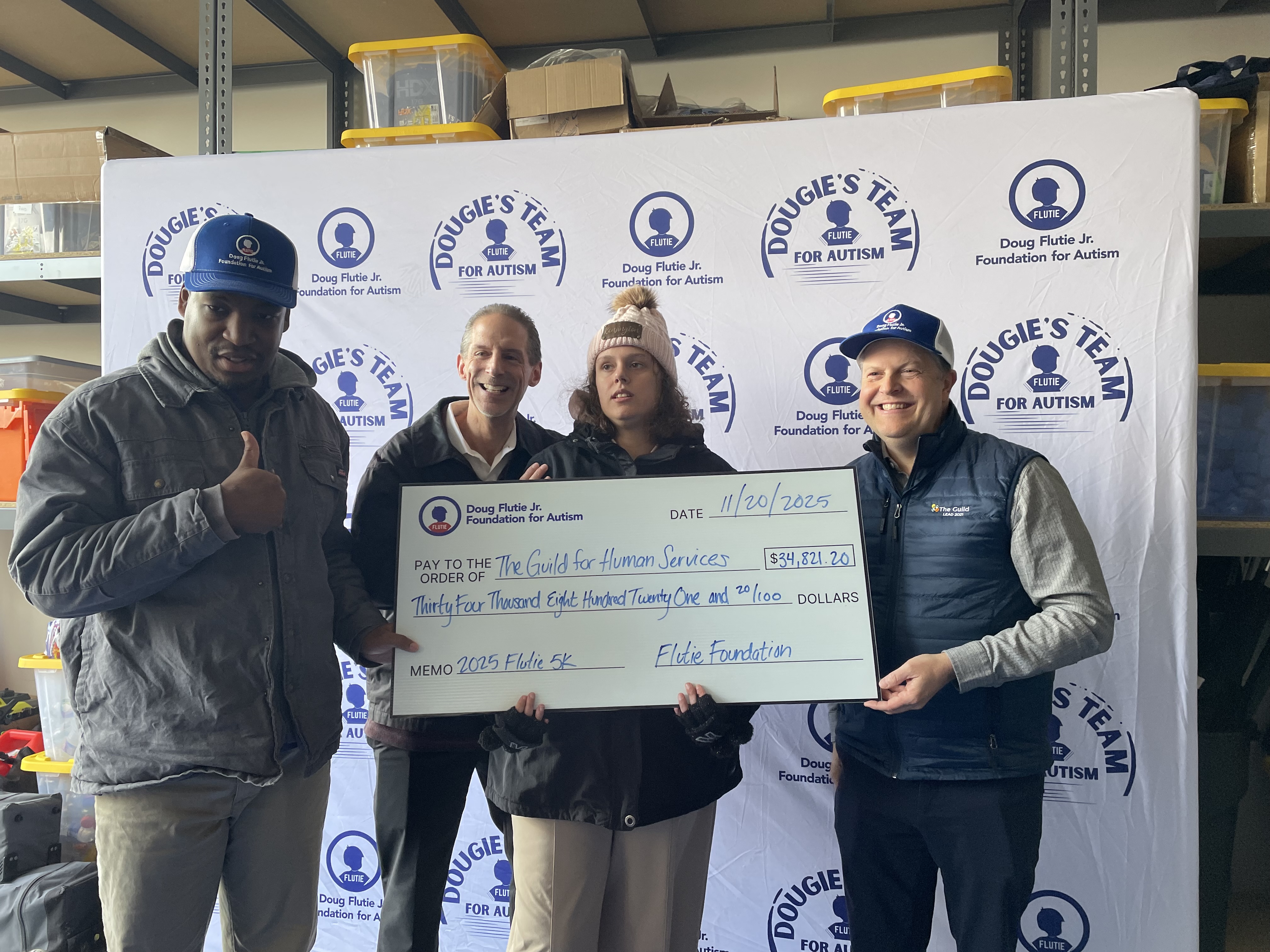 A group of people representing The Guild hold up an oversized fundraising check from The Doug Flutie Foundation in front of a photo backdrop with The Flutie 5K logo.