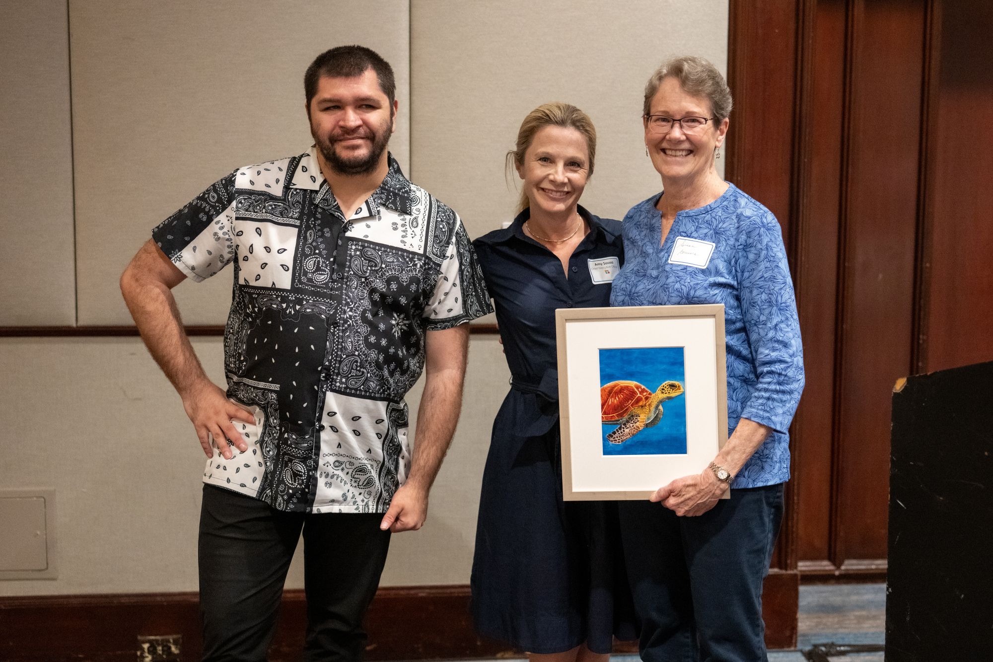 Dr. Susan Connors holds an original artwork of a sea turtle, standing to the right of an adult resident and CEO Amy Sousa.
