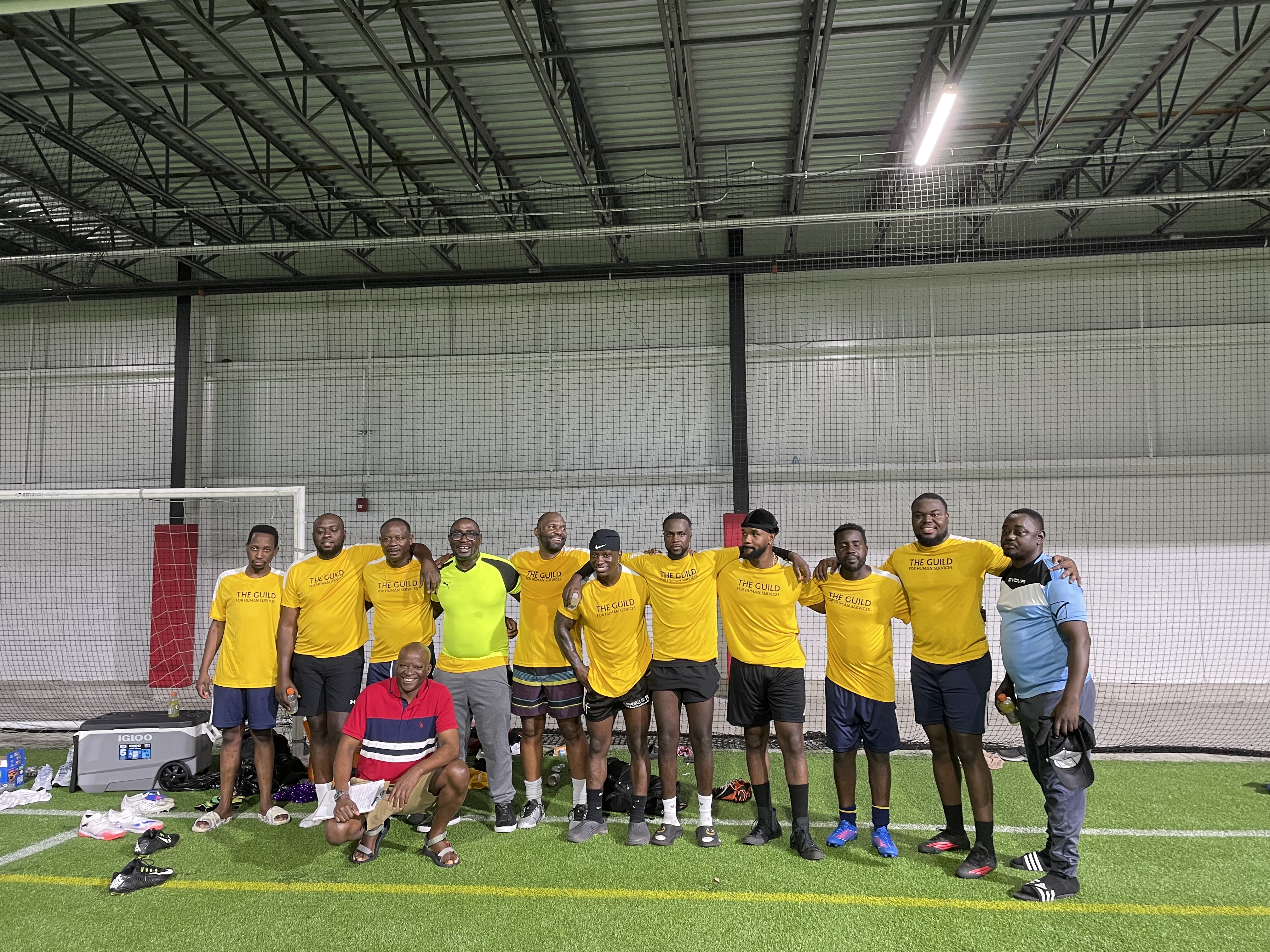 A group of men stand together in front of a goal on an indoor soccer field with their arms around each other wearing matching yellow soccer jerseys.