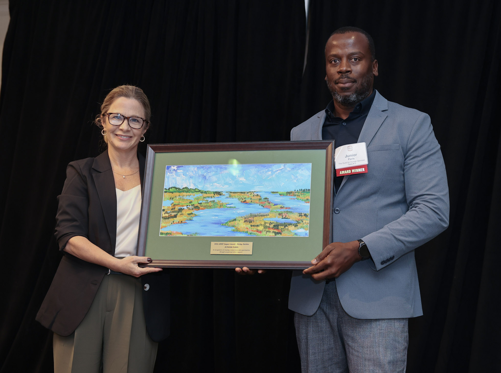 Amy Sousa and Junior Paris stand together in front of a black backdrop, each holding one side of a framed painting of water and marshland.
