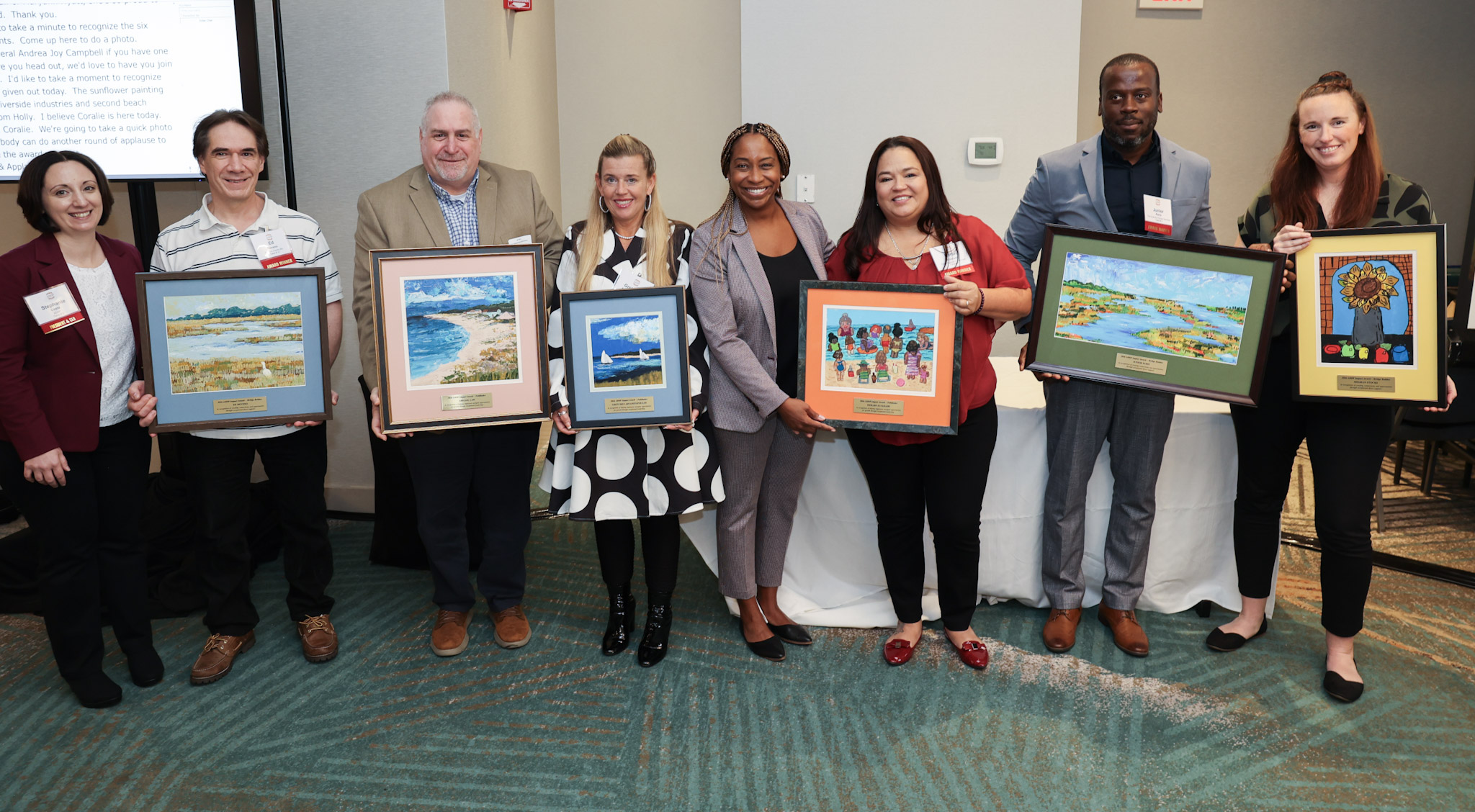 An ADDP staff member and the Massachusetts Attorney General Andrea Campbell stand together with the six award recipients and their framed paintings in a conference room.