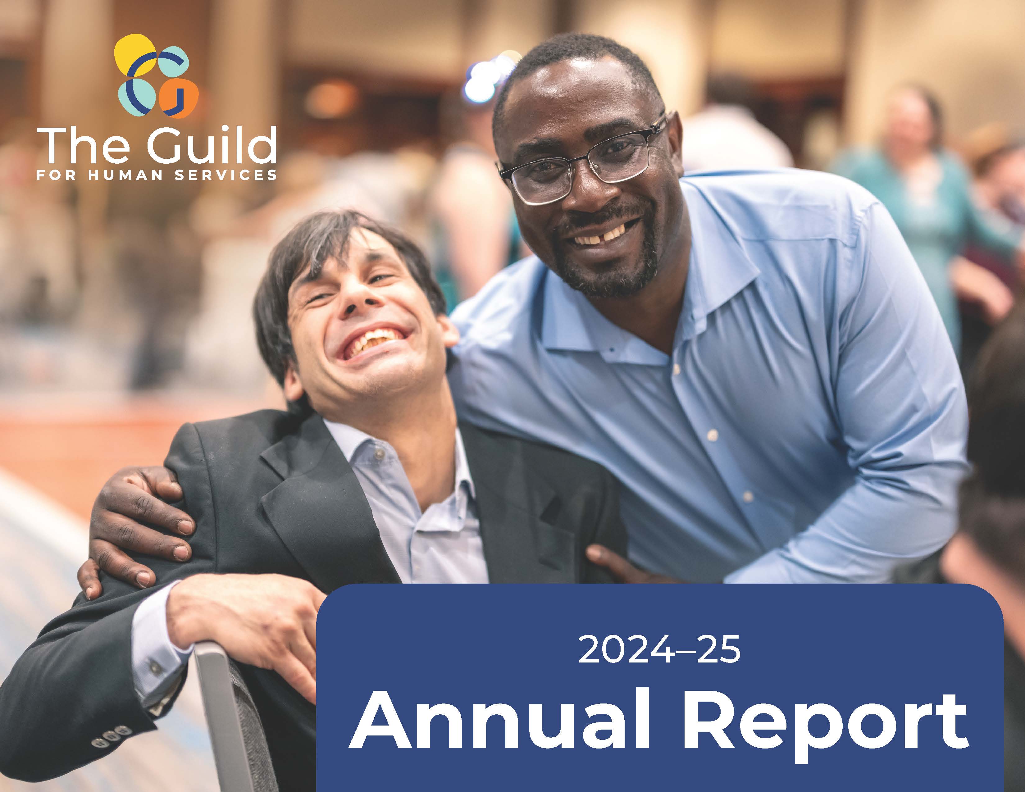 A staff member puts his arm around an adult resident sitting sideways in a chair as they both smile widely.