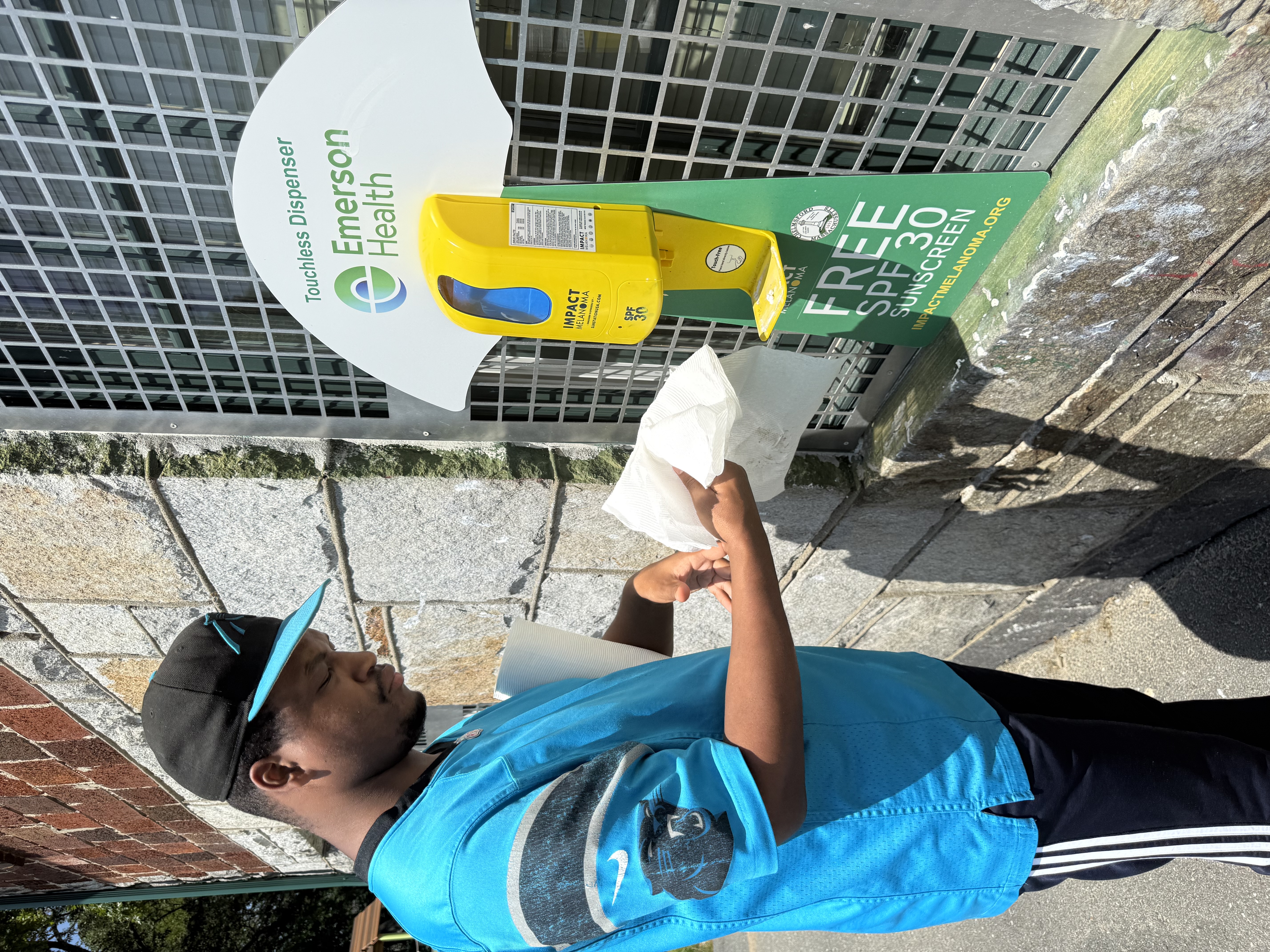 An adult resident holds a paper towel next to a bright yellow touchless sunscreen dispenser out in the community.