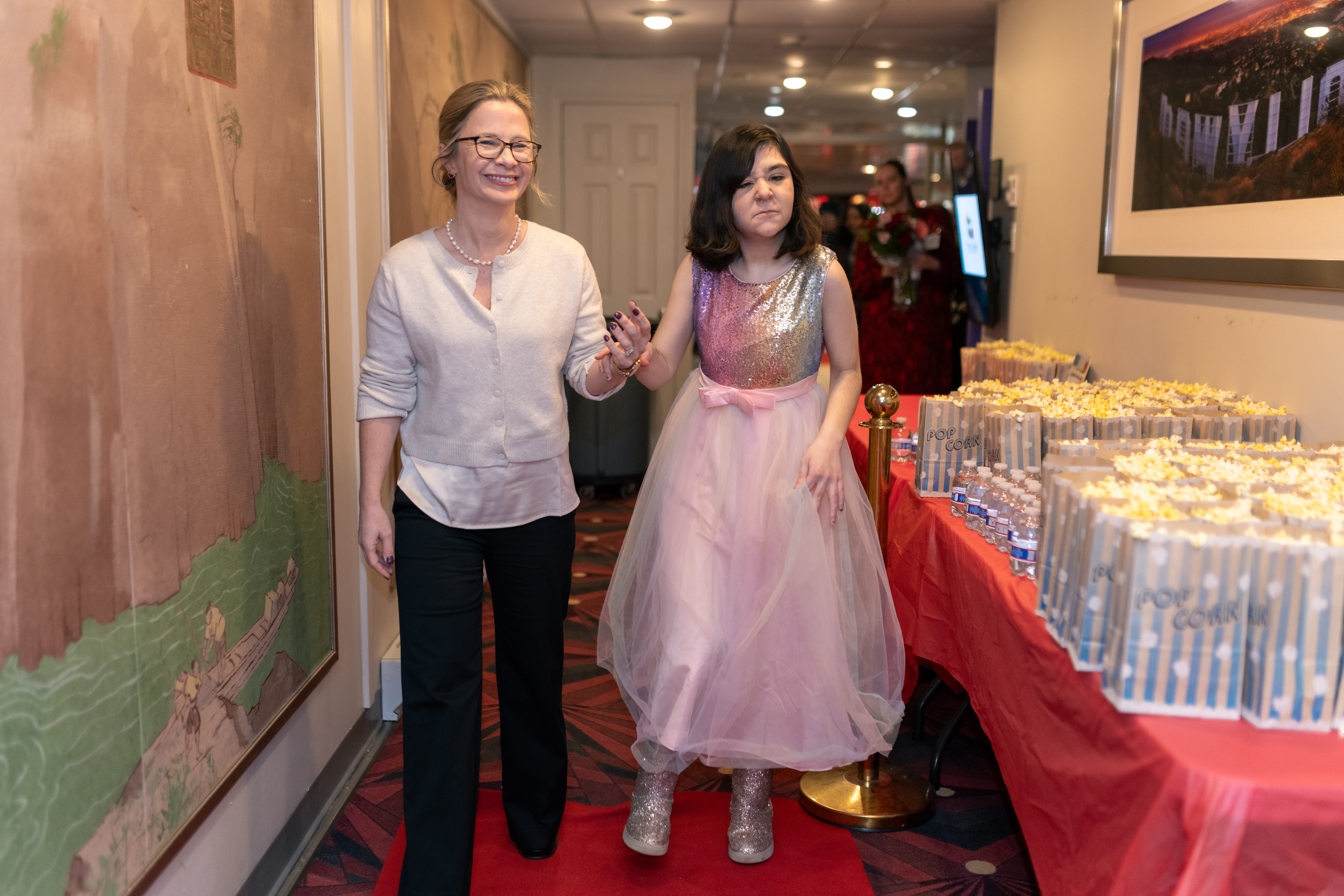 CEO Amy Sousa, in black slacks and a cream-colored sweater, and documentary star Kaitlyn, in a pink dress with a sequined top and matching sequined boots, walk down a red carpet with bags of popcorn lining a table on their right.