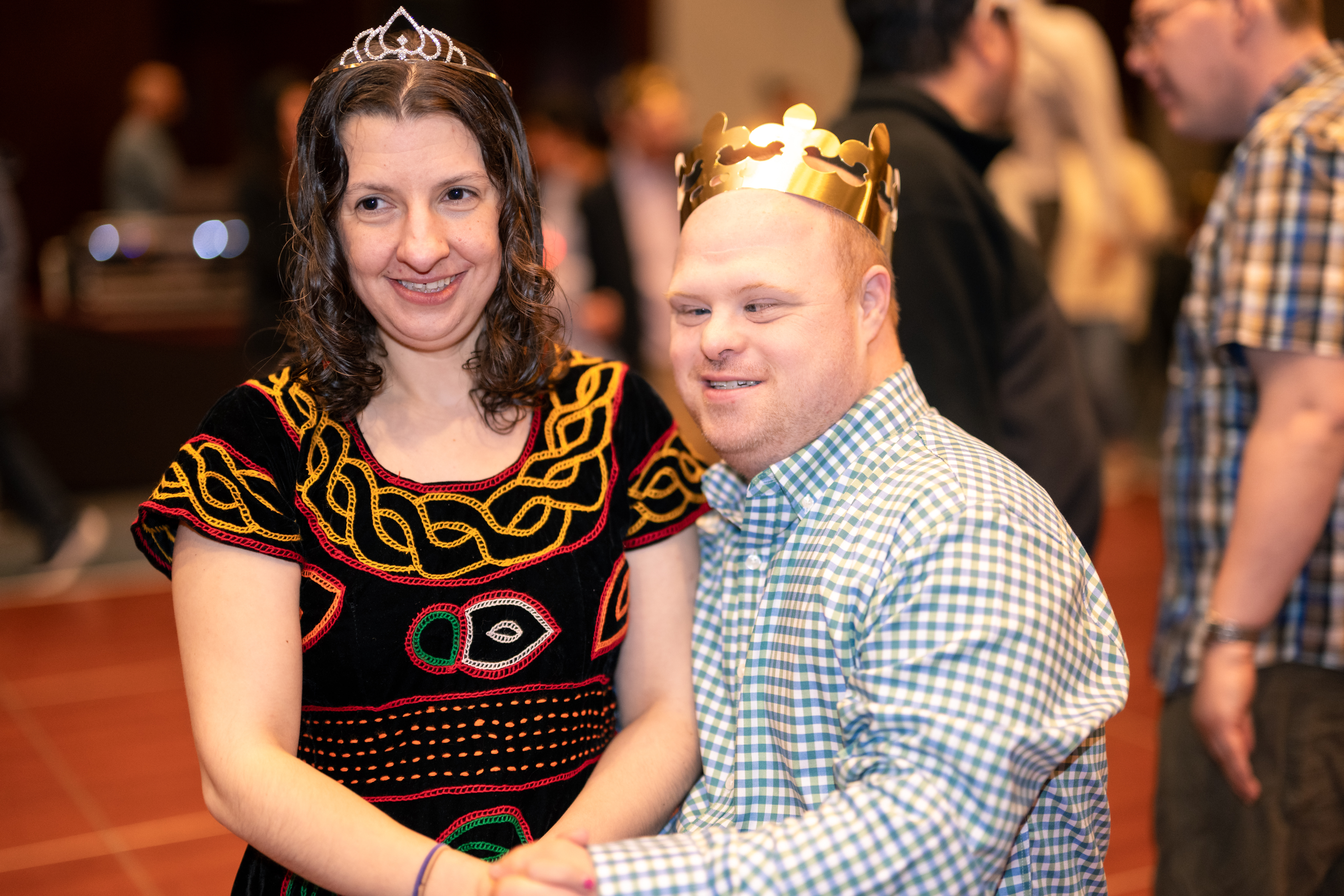 Two adult residents from The Guild stand smiling arm-in-arm on the dance floor with people behind them and wearing gold crowns.