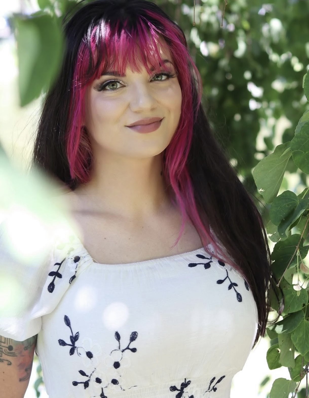 Kaitlyn Foehl stands outside among green tree branches wearing a white top with black floral designs.