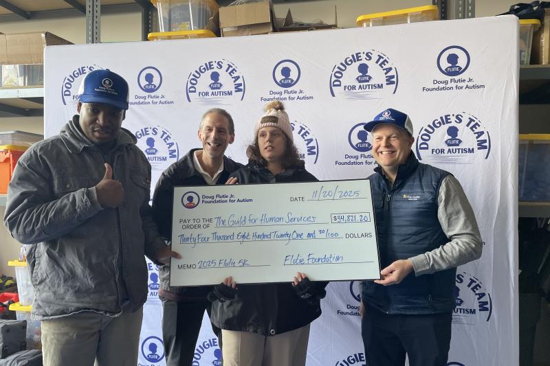 A group of people representing The Guild hold up an oversized fundraising check from The Doug Flutie Foundation in front of a white photo backdrop with the Flutie 5K logo repeated.