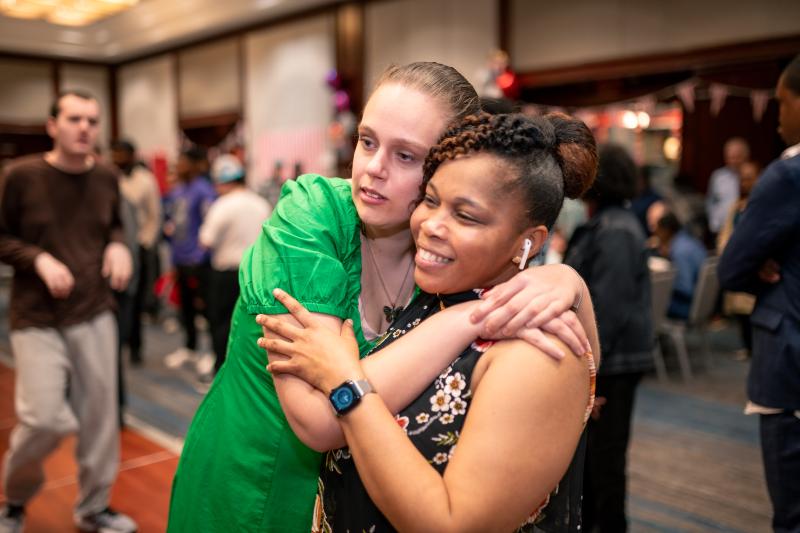 An adult resident in a green dress hugs a staff member in the ballroom of a busy event.