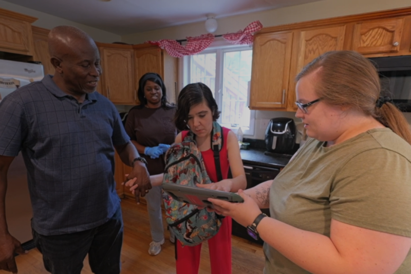 A woman stands in the kitchen of her home wearing a backpack and reaching to use her communication device. Staff are standing with her and one staff member is holding the device.