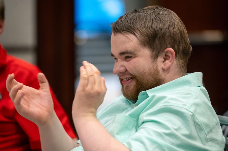 An adult resident sits at a table and smiles while clapping, looking off camera.