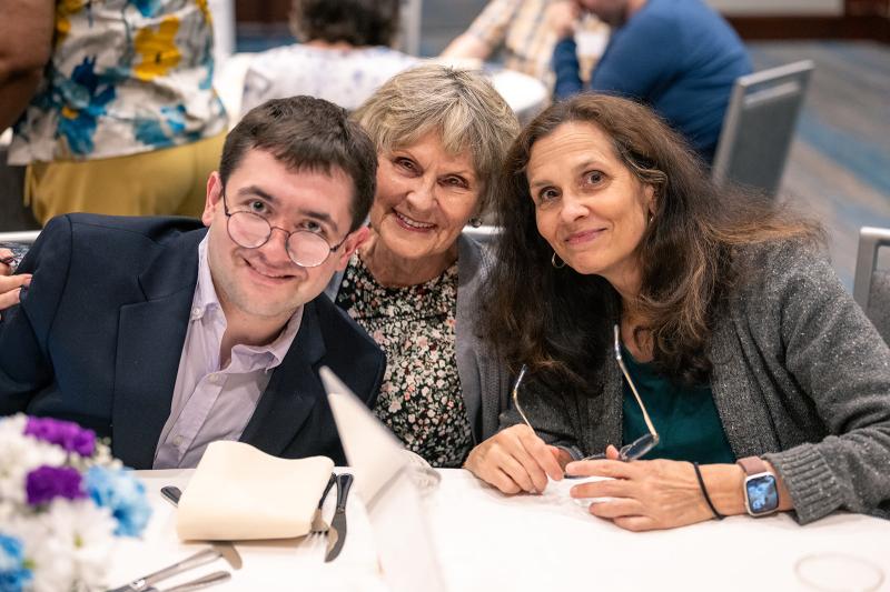 An adult resident wears a suit jacket and leans in close with two family members while seated together at a table.