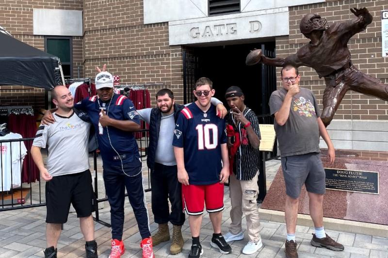 Six adult residents stand in a line with their arms around each other in front of a statue of a football player.