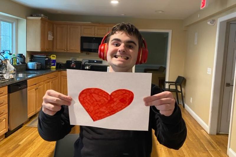 A student stands in the kitchen of his residence and holds up a hand-drawn red heart on a white piece of paper.
