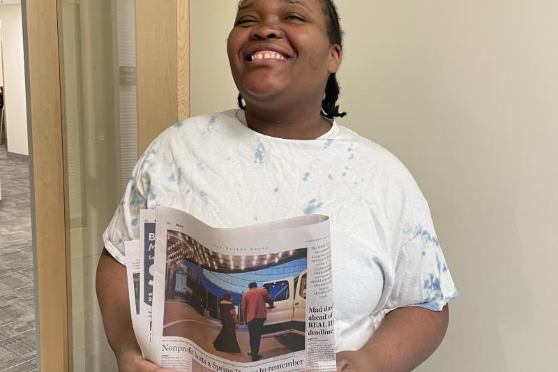A woman stands in an office doorway and holds up a copy of The Boston Globe featuring an image of her on the front page.