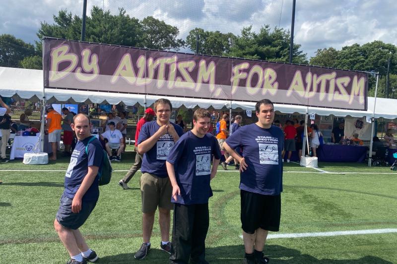 A group of four adult residents stand in a line wearing matching t-shirts in front of a banner reading "By autism, for autism"