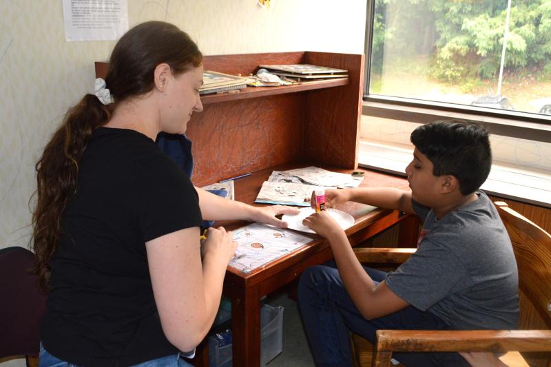 A student sits at a desk with papers while a staff member sits to the side working with him.