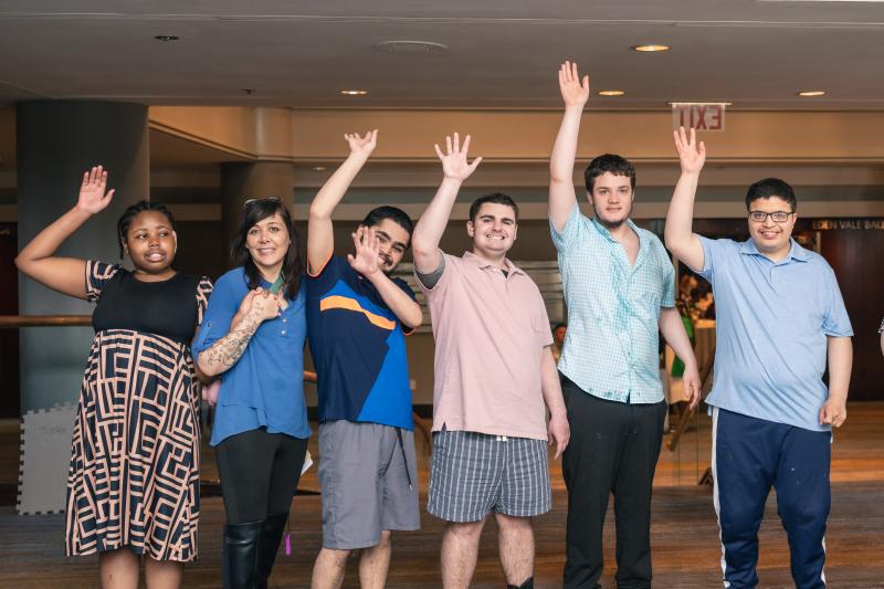 A group of five students and their teacher stand together with their hands raised to wave at the camera.