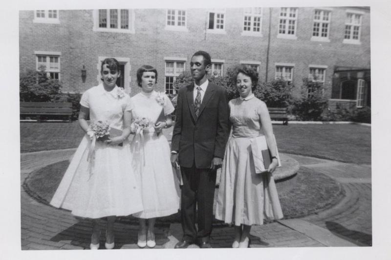 A black and white photo depicts four adults standing together on a lawn outside of a building.