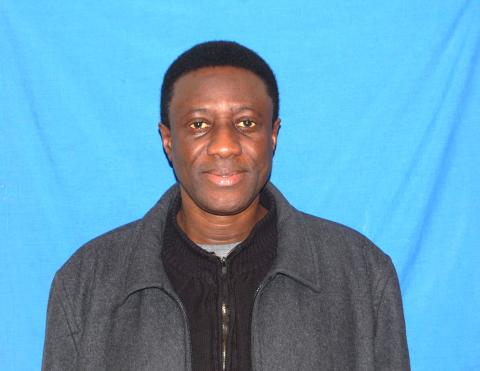 Alpha stands in front of a plain blue background and looks into the camera with a neutral expression.