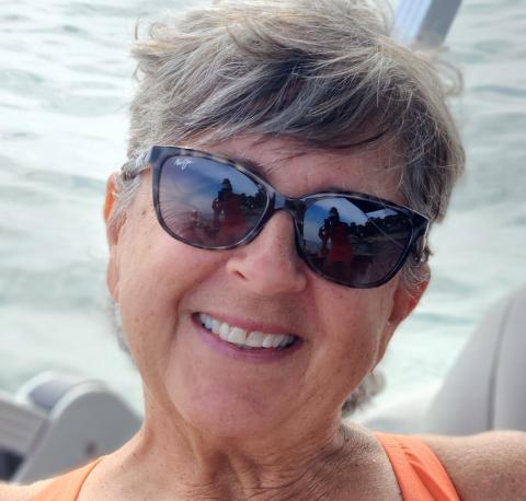 A close up image shows Rose in front of a body of water wearing sunglasses and smiling.