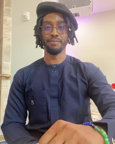 Uzoma sits with a hand on a table and looks straight on into the camera with a neutral expression.