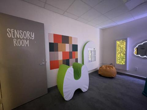 One view of a sensory room shows felt tiles on the wall, a curved compression chair, a bean bag in the corner, two interactive panels with different lights, and a cloud-shaped mirror.