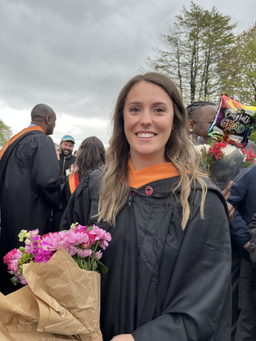 Morgan stands outside on a cloudy day in a black graduation gown, holding a boquet of flowers.