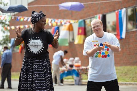 A woman in a Crocodile River Music t-shirt leads a man in a dance outside in front of colorful banners representing flags from around the world.