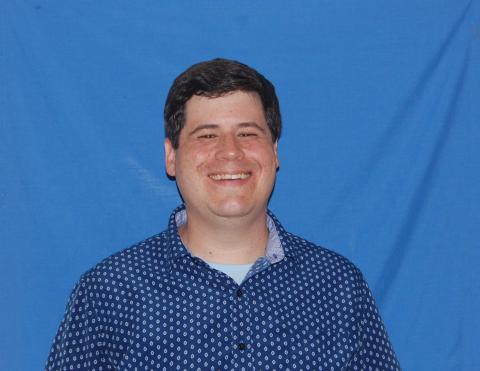 Chad stands smiling in front of a plain blue background.