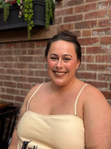 Tori Carey sits in front of a brick wall and smiles at the camera wearing a pastel yellow shirt.