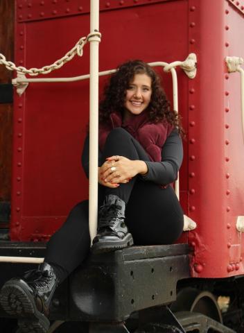 Cassie May sits in front of a red train car wearing a red vest and smiling at the camera.