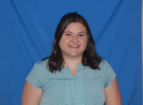 Kirstin stands against a plain blue background and smiles, wearing a light blue shirt with a small pattern in darker blue.