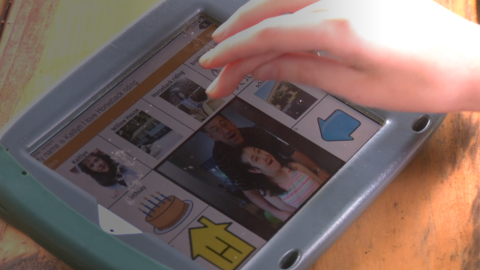 A hand hovers over a "horseback riding" icon on a close-up shot of a speech-generating device on a table.