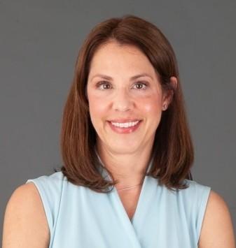 Mary Giunta is smiling directly into the camera and wearing a light blue shirt in front of a medium gray background.