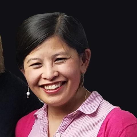 Oanh Bui smiles while wearing a bright pink shirt and cardigan and sitting slightly sideways in front of an all-black background.