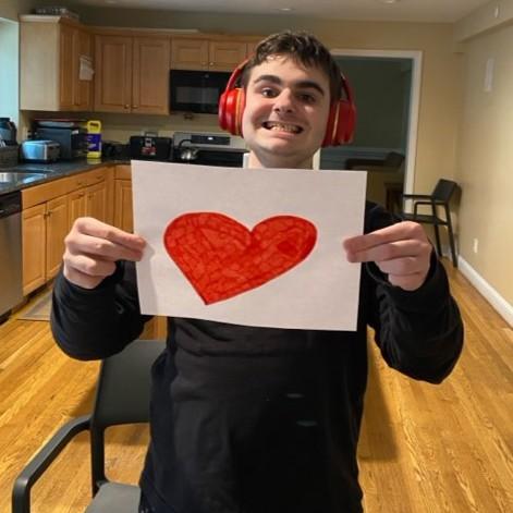 A student wears red noise-canceling headphones and smiles in his house while holding up a hand-drawn red heart on a white piece of paper.