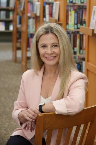 Amy Sousa headshot - she wears a light pink blazer and sits in a chair in front of bookcases, smiling at the camera.