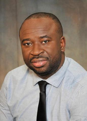 Jerry Yahaya sits in front of a gray backdrop wearing a light blue shirt and a black tie.