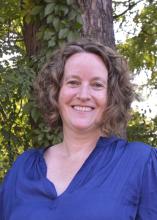 Katharine stands in the shade beneath a tree wearing a bright blue blouse and smiling into the camera.