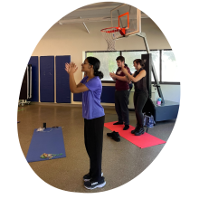 Akila Muthukumar stands to the side of a yoga mat and crosses one arm over the other. A student and teacher can be seen following her movements in the background.