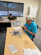Two adult residents sit at conference room tables at The Guild offices with piles of IMPACT Melanoma materials they are working on putting into packets.