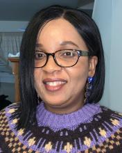 Nerline wears a purple and black woven sweater and glasses and smiles into the camera.