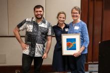 Dr. Susan Connors holds an original artwork of a sea turtle, standing to the right of an adult resident and CEO Amy Sousa.